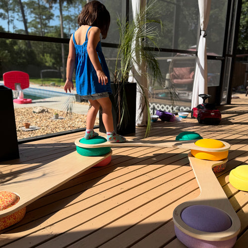 A child playing on a wooden balance board with colorful stepping stones in the background on a wooden deck.