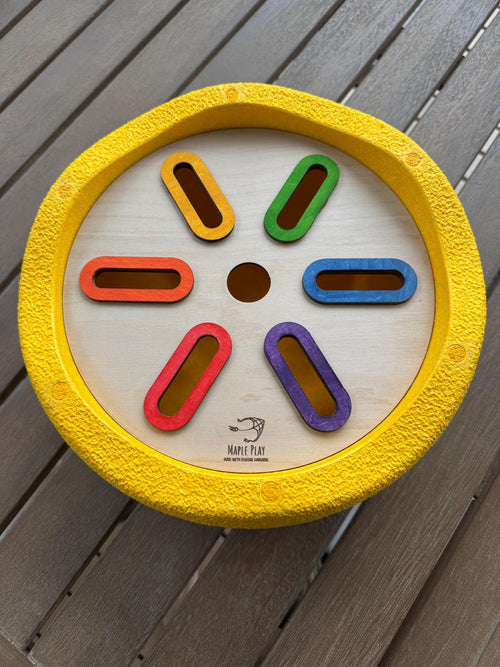 A wooden sorting insert with colorful elongated slots, designed for use with Stapelstein stepping stones. The insert has a yellow border and is displayed on a wooden surface.