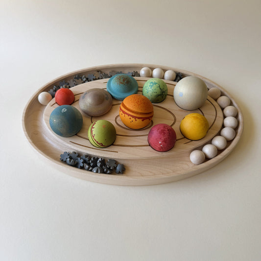 A wooden solar system model toy set on a light wood tray, featuring colorful painted planets, a central sun, and small dark asteroids arranged on a plain cream background.